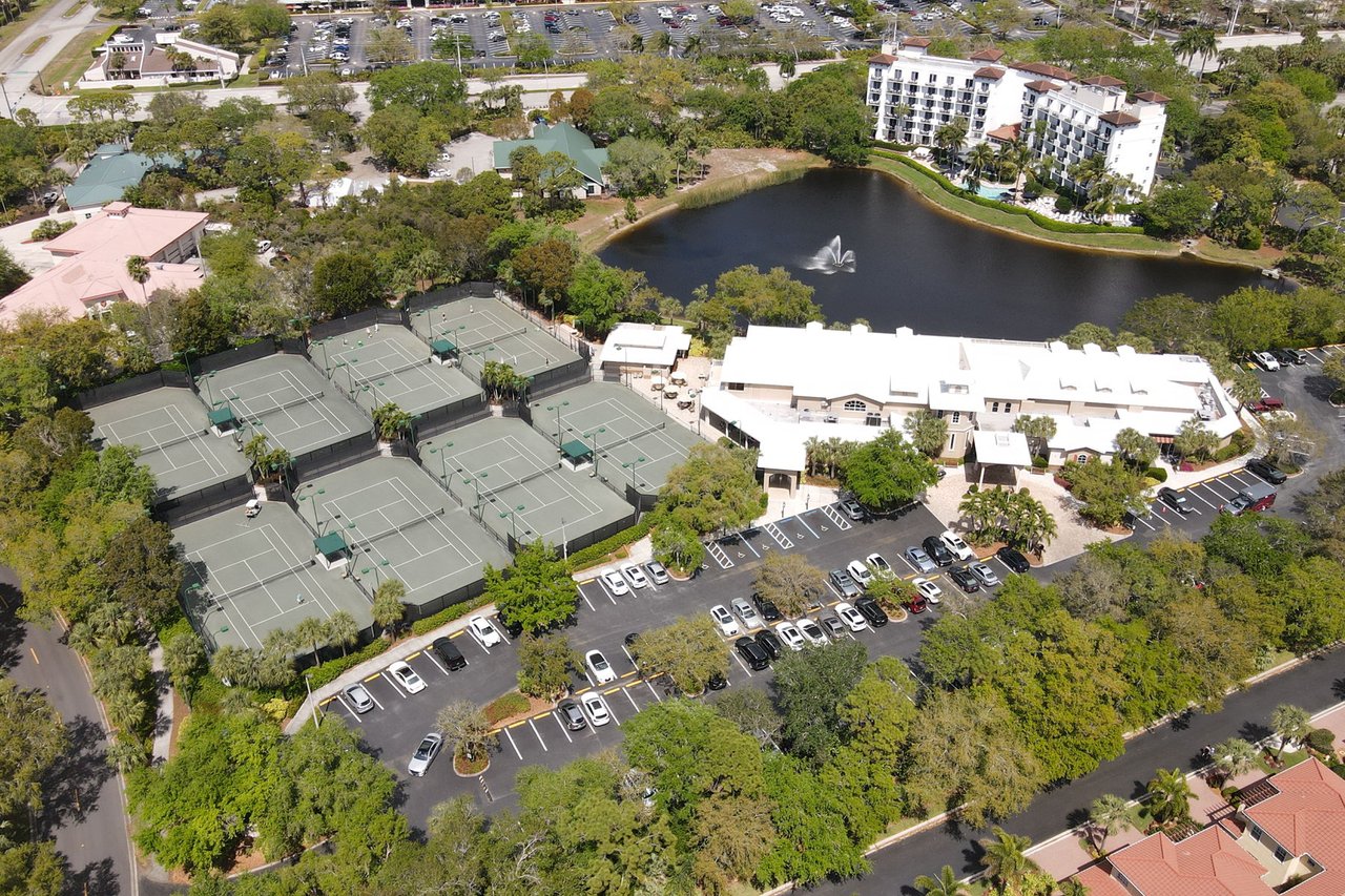 Aerial view of Pelican Bay tennis courts beside a clubhouse and pond with a fountain.