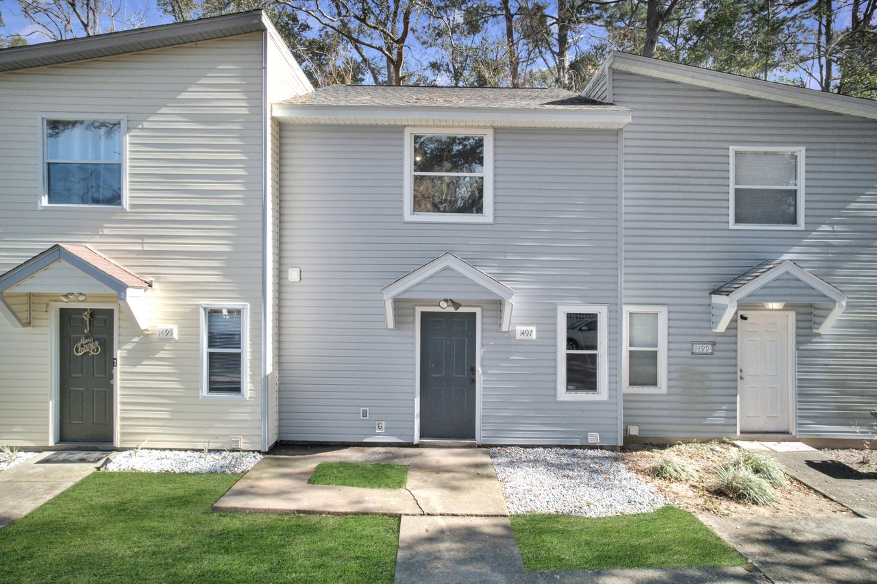 Row of three townhouses with gray siding and small front porches. Well-maintained lawns and trees in the background under a clear sky.