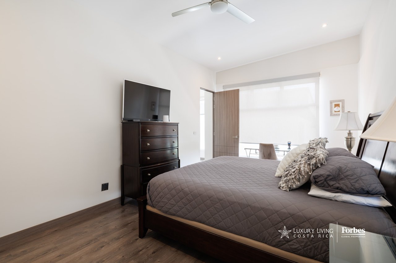 Furnished bedroom with a gray quilted bed, dark wood dresser, and television beside a bright window.