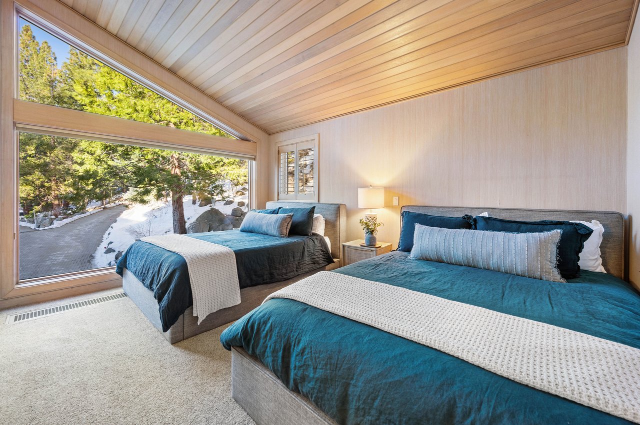 Bedroom with two teal-dressed beds, a vaulted wood ceiling, and a large window overlooking snowy evergreens.