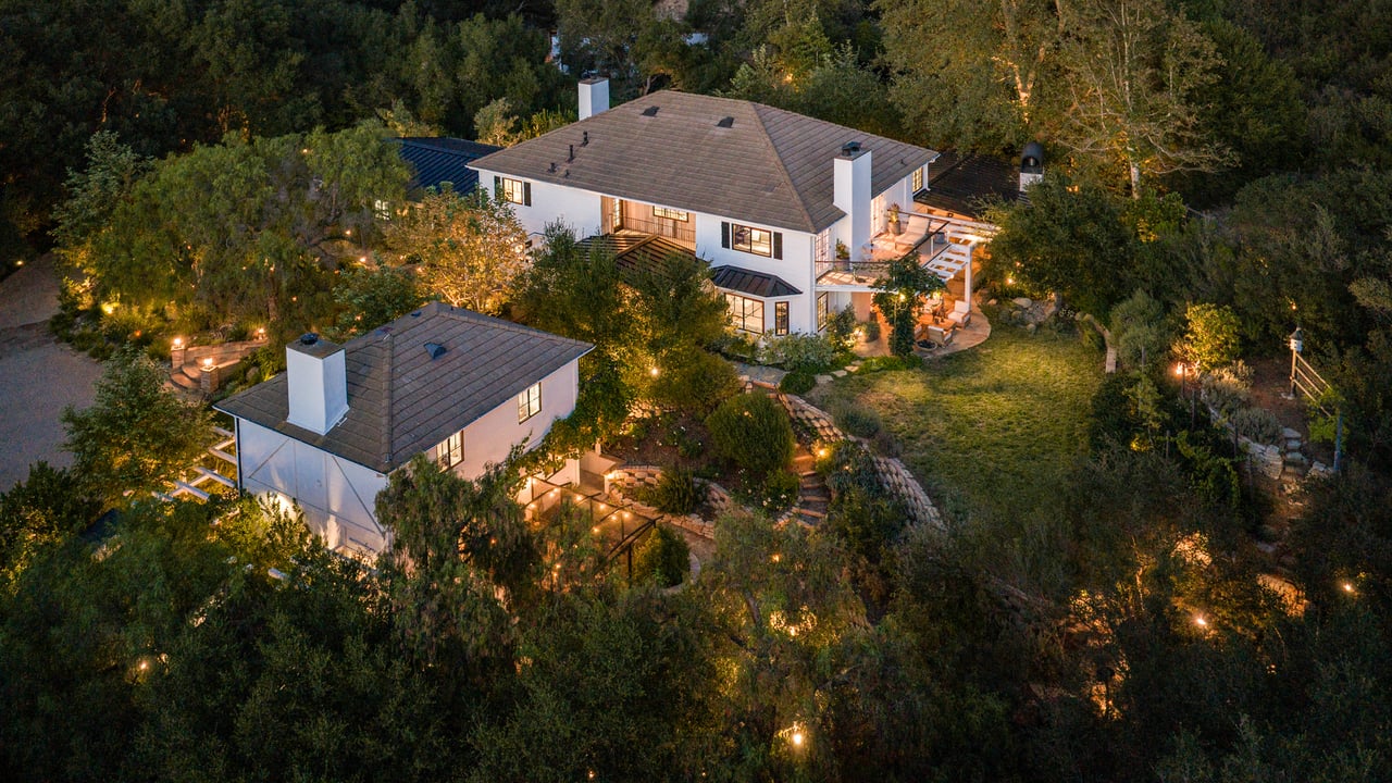 Aerial view of a white home and detached outbuilding surrounded by trees at dusk.