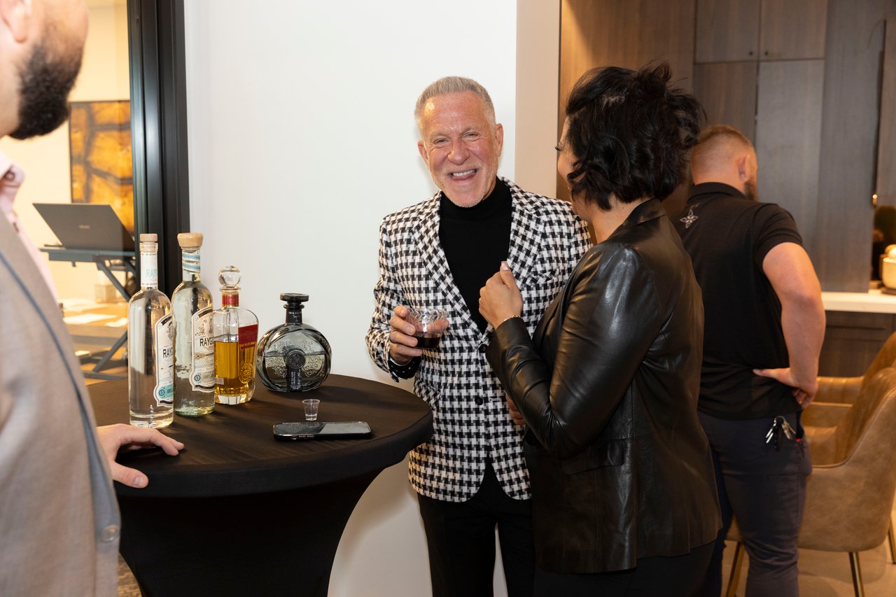 Guests mingle at an open house beside a table of drinks; a man in a checked blazer chats with a woman.