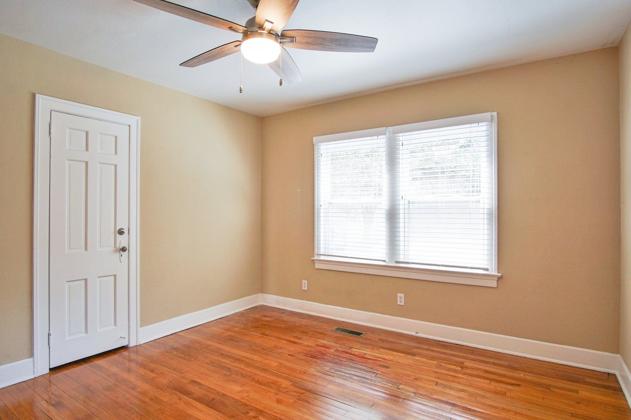 An empty room with beige walls and polished wooden floors. A ceiling fan is on above. Sunlight streams through a window with blinds, creating a bright, airy feel.