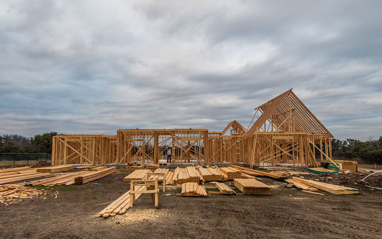 House under construction with exposed wooden framing and lumber stacked across the dirt lot.