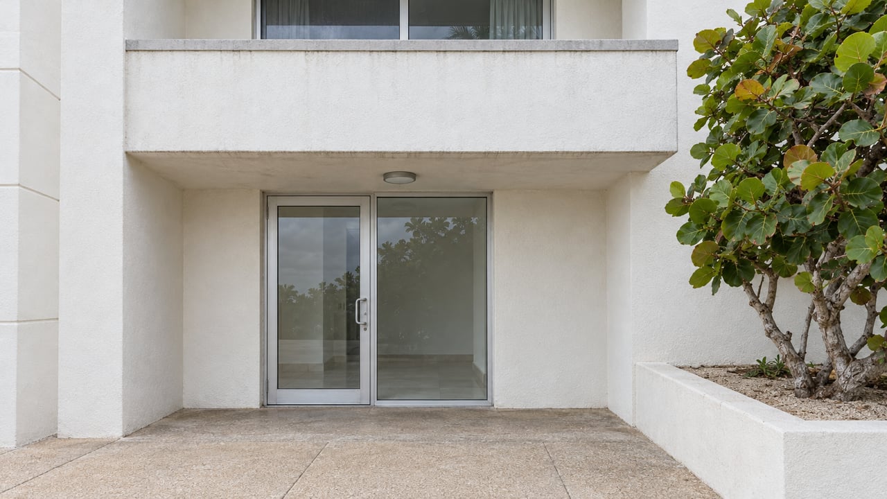 White rendered condominium entrance with closed glass doors, a concrete balcony and a sea grape tree in a raised planter.
