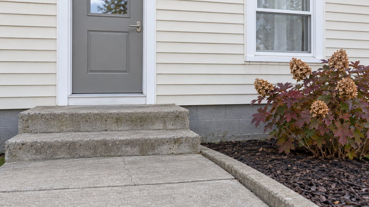 Concrete front stoop and gray entry door beside cream siding and an oakleaf hydrangea with autumn foliage.
