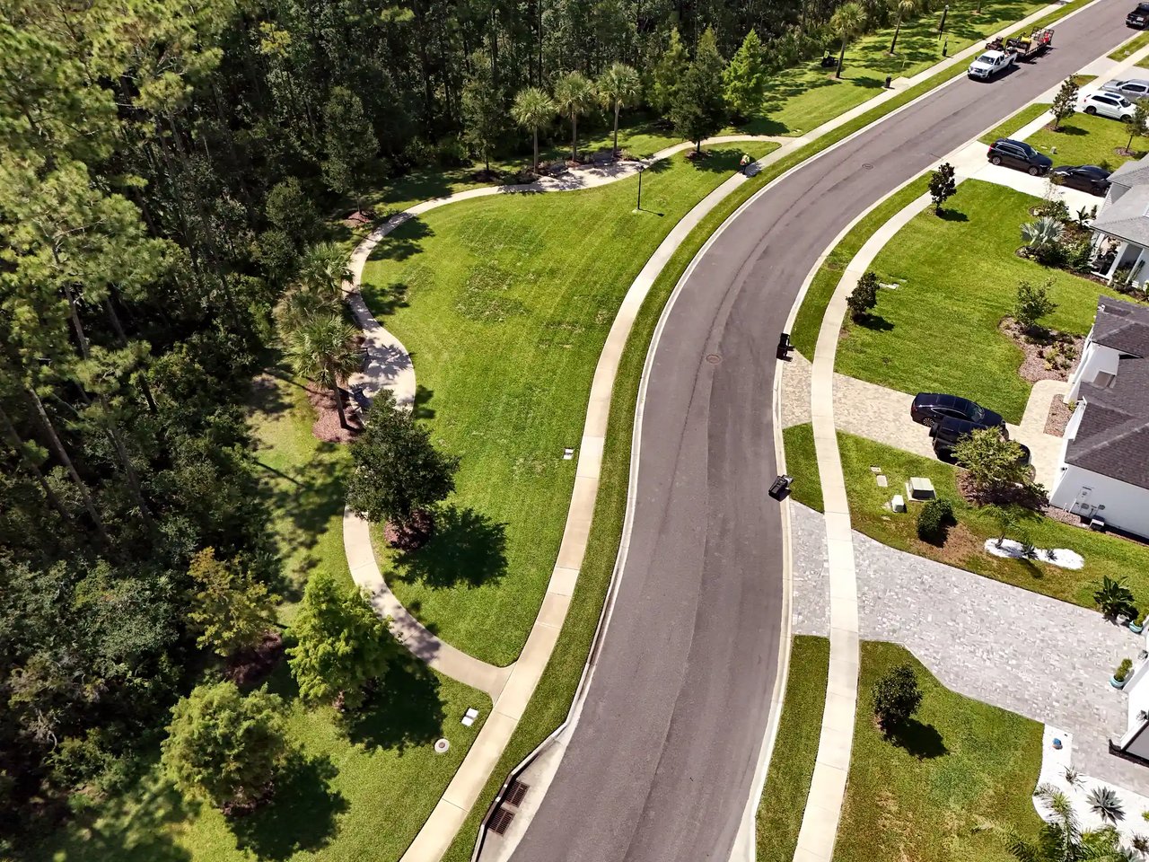 Aerial view of a curving walking path around an open green space beside a Palm Crest street, bordered by pines.