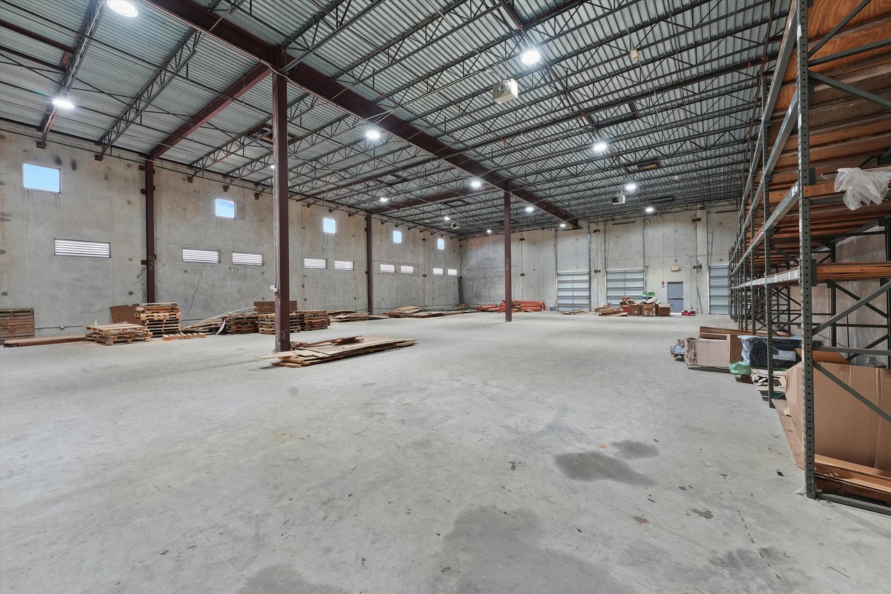 Large warehouse interior with high metal-truss ceiling, open concrete floor, loading doors, and perimeter storage racks.