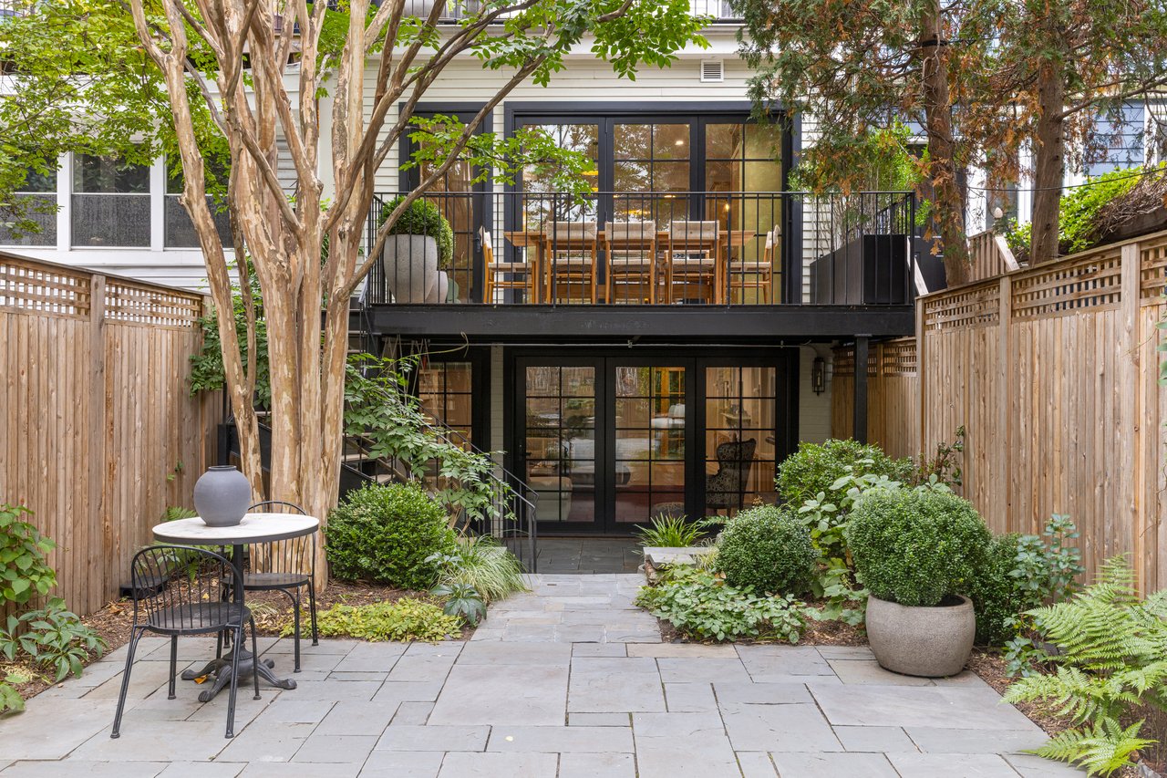 Rear exterior view of a townhome featuring a black metal deck with an outdoor dining area, overlooking a lush stone patio garden with privacy fencing.