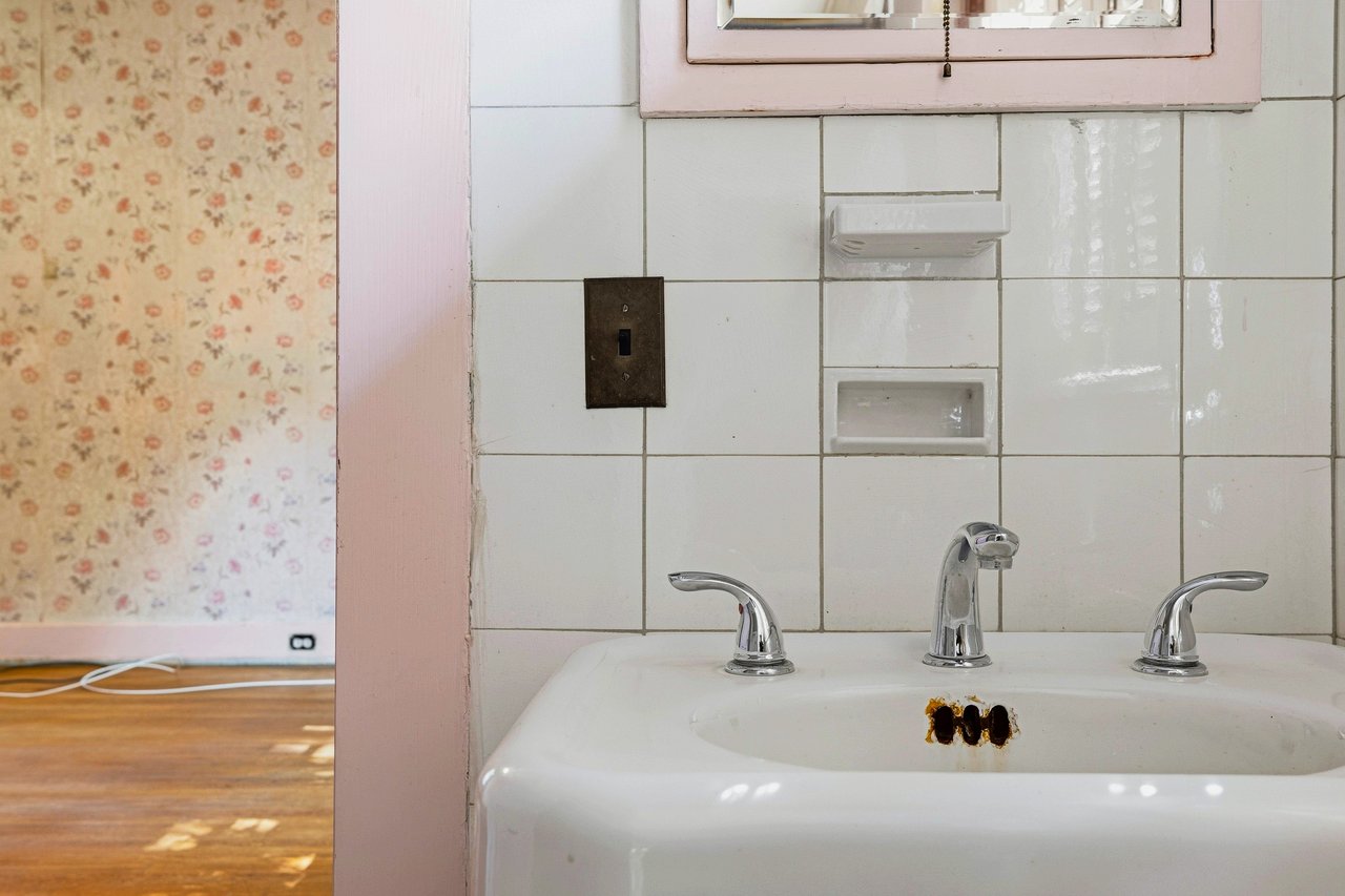 Bathroom sink with chrome faucets, white tile, pink trim, and floral wallpaper visible through the doorway.