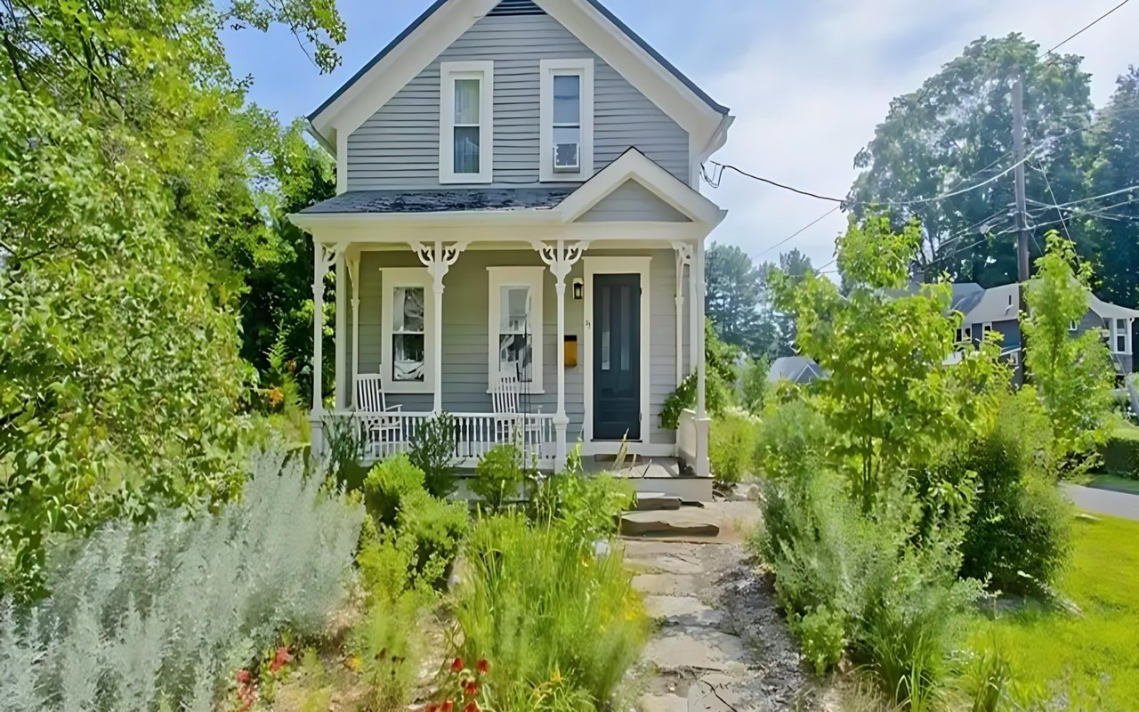 Gray farmhouse with a white-trimmed porch, leafy garden, and stepping-stone path to the entrance.