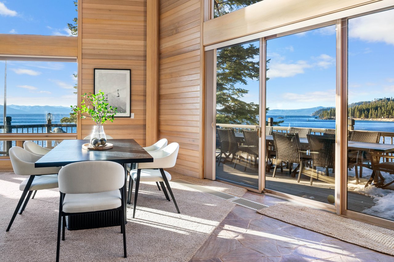 Lakefront dining room with wood-paneled walls, white chairs, and sliding doors opening to a deck with mountain views.