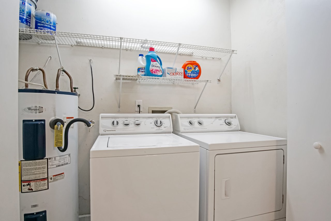 Laundry room with a washer, dryer, and water heater. Shelves above hold detergent and fabric softener. The space is organized and functional.