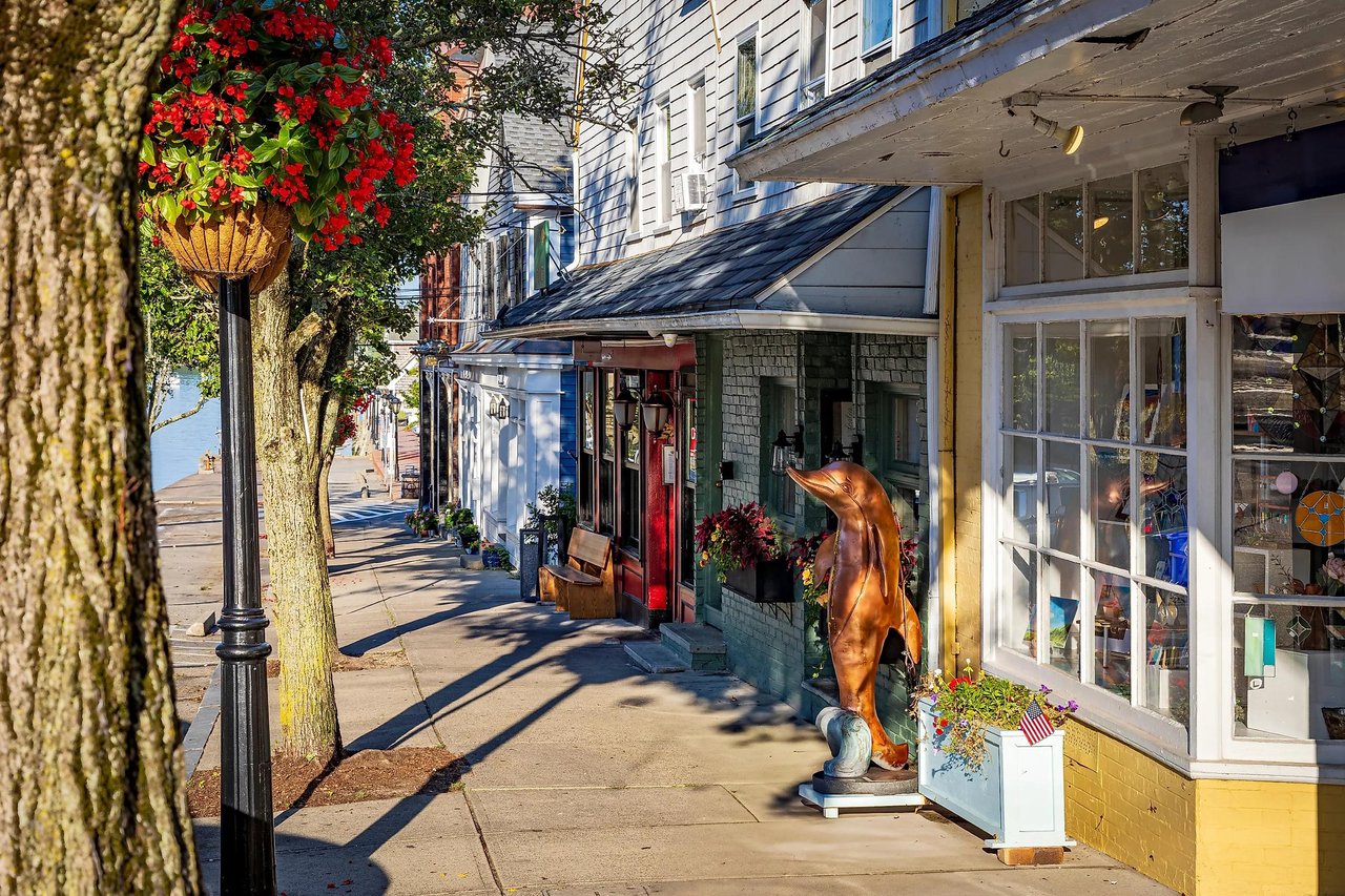 Small-town storefronts along a sunny sidewalk, with hanging flowers and a copper dolphin sculpture.
