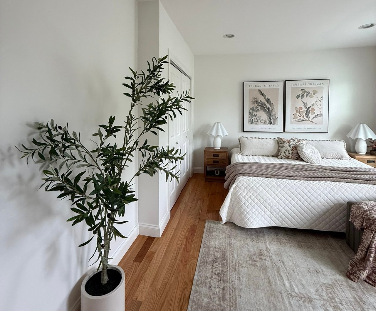 Bright bedroom with a quilted bed, botanical prints, wood floors, and a potted tree.