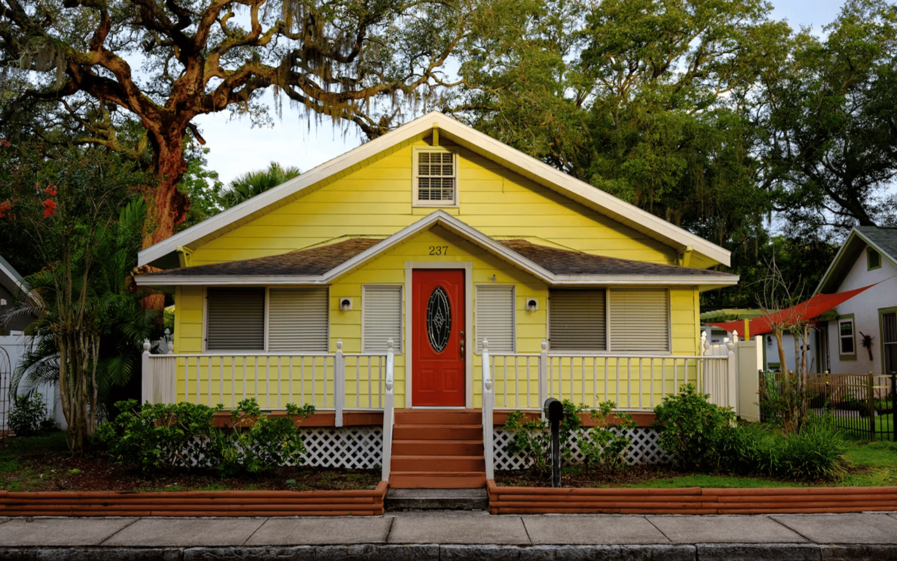Yellow house with a red front door and white porch beneath sprawling oak trees.