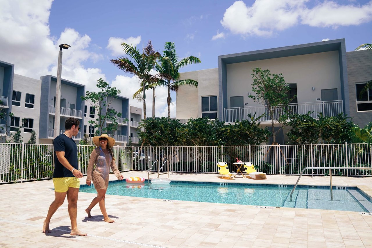 Residential courtyard pool with a tiled deck, yellow lounge chairs, palm trees, and modern low-rise buildings.