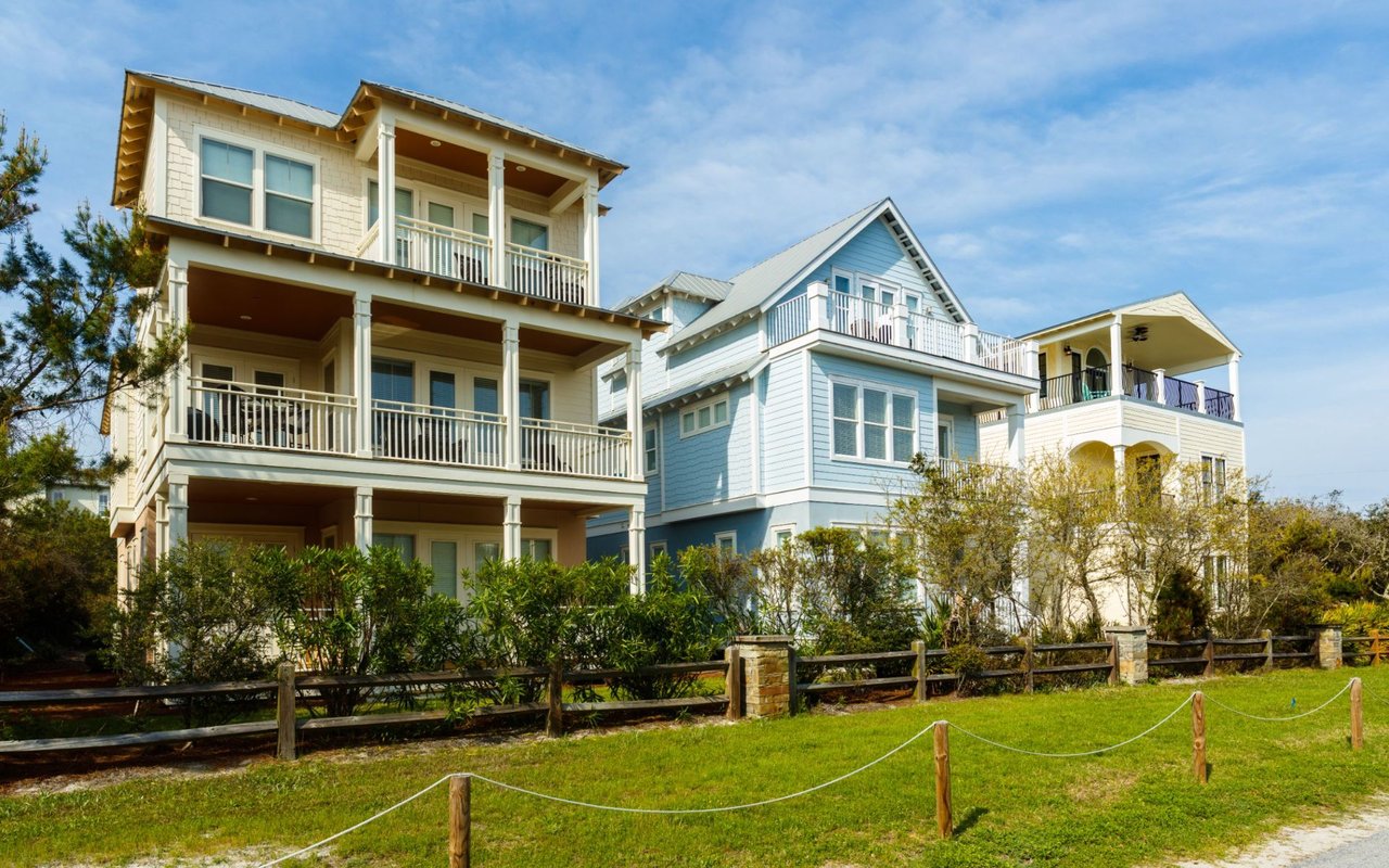 Three coastal-style homes with balconies behind a grassy lawn and wooden fence.