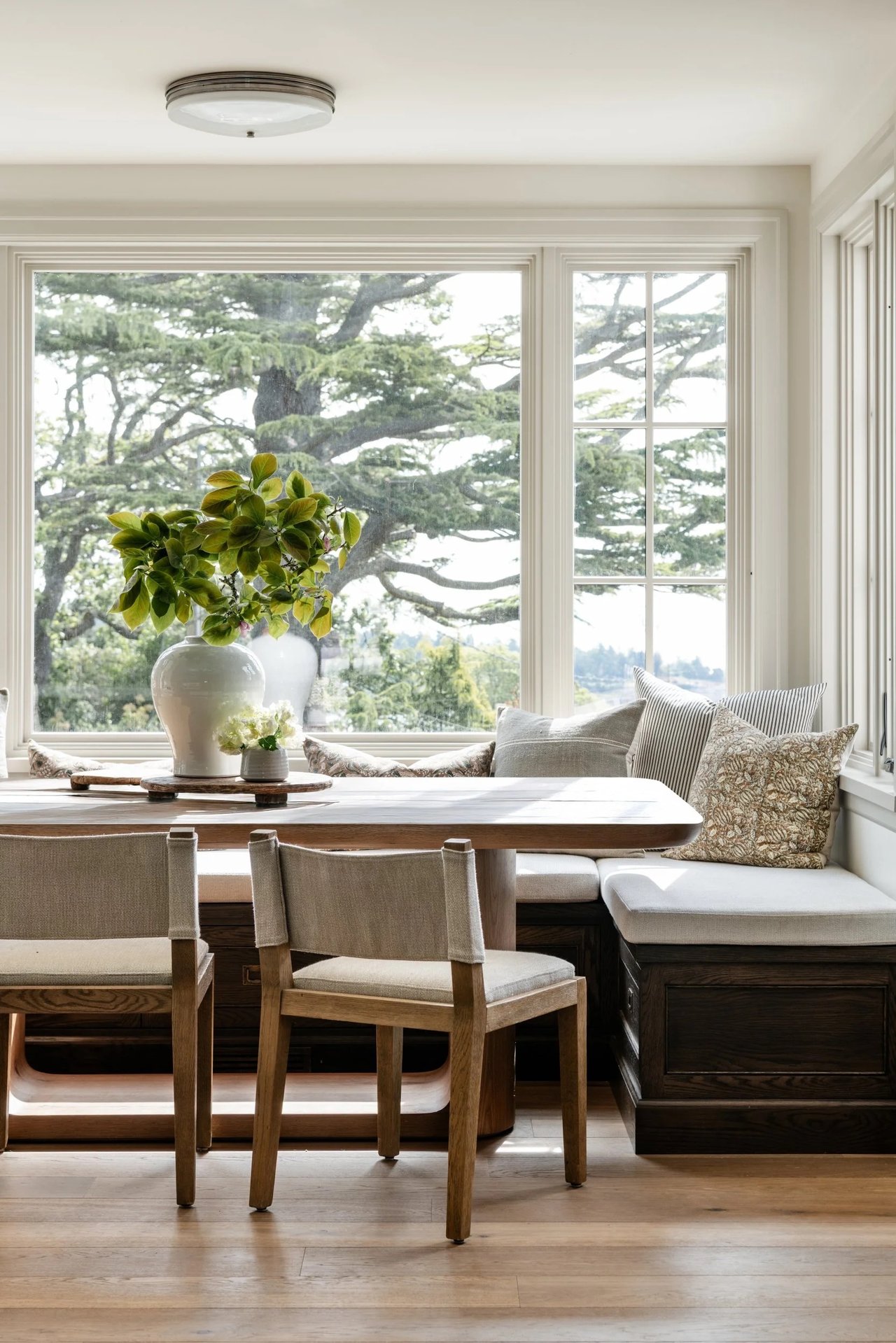 Bright dining nook with a wooden table, cushioned banquette, and large windows overlooking trees.