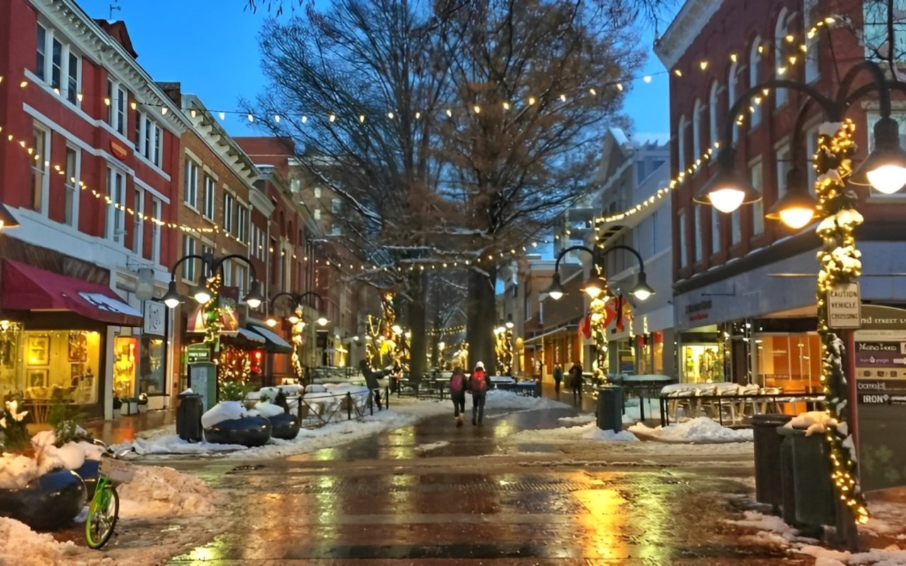 Snowy downtown street lined with brick shops and glowing holiday lights at dusk.
