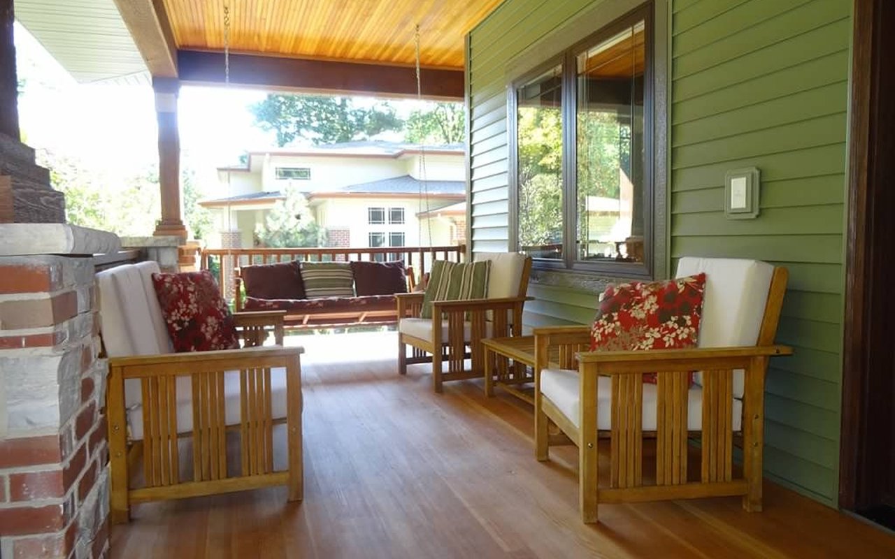 Covered porch with wooden seating, floral pillows, and a brick-and-stone fireplace.