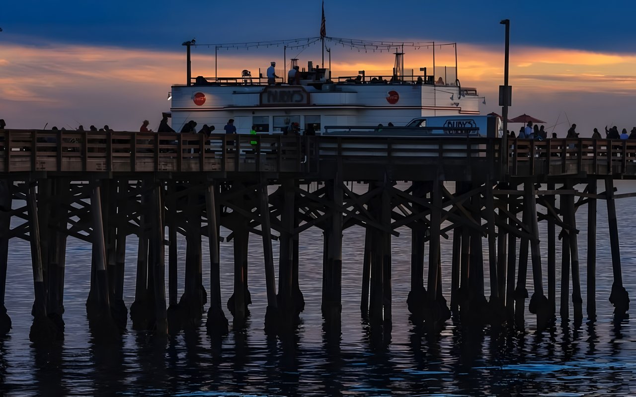 A pier restaurant with people fishing on it, silhouetted against a colorful sunset over the ocean.