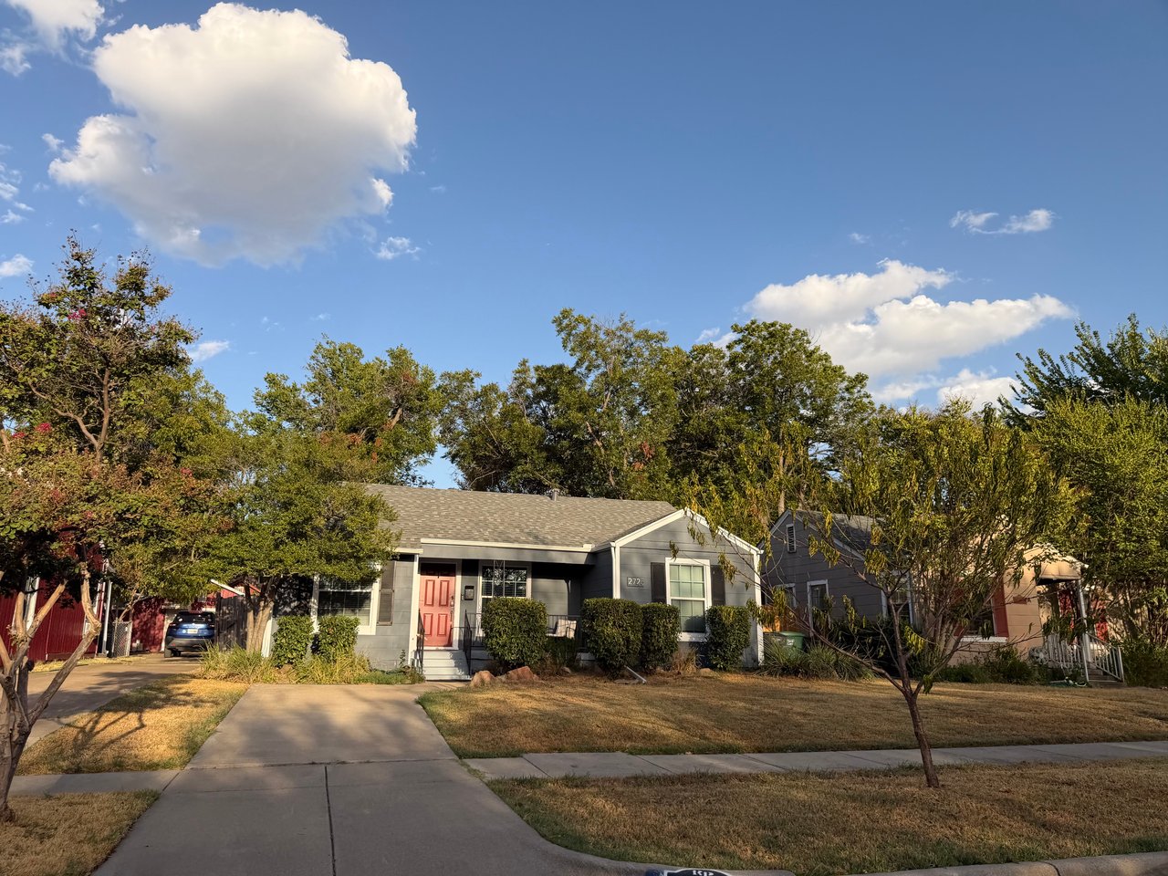 Gray single-story home with a red front door, driveway, and mature trees.