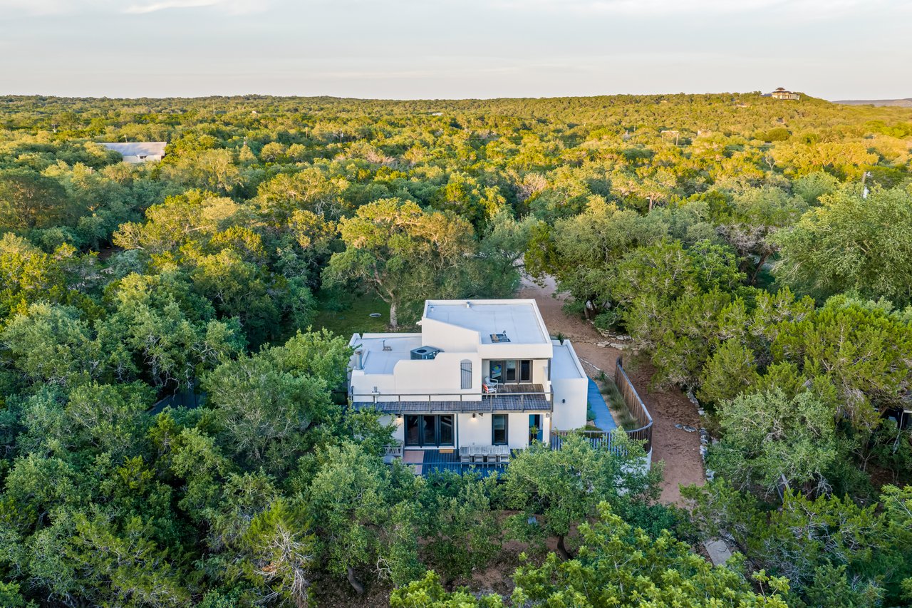 Aerial view of a modern white home nestled among dense trees, with a small pool and deck.