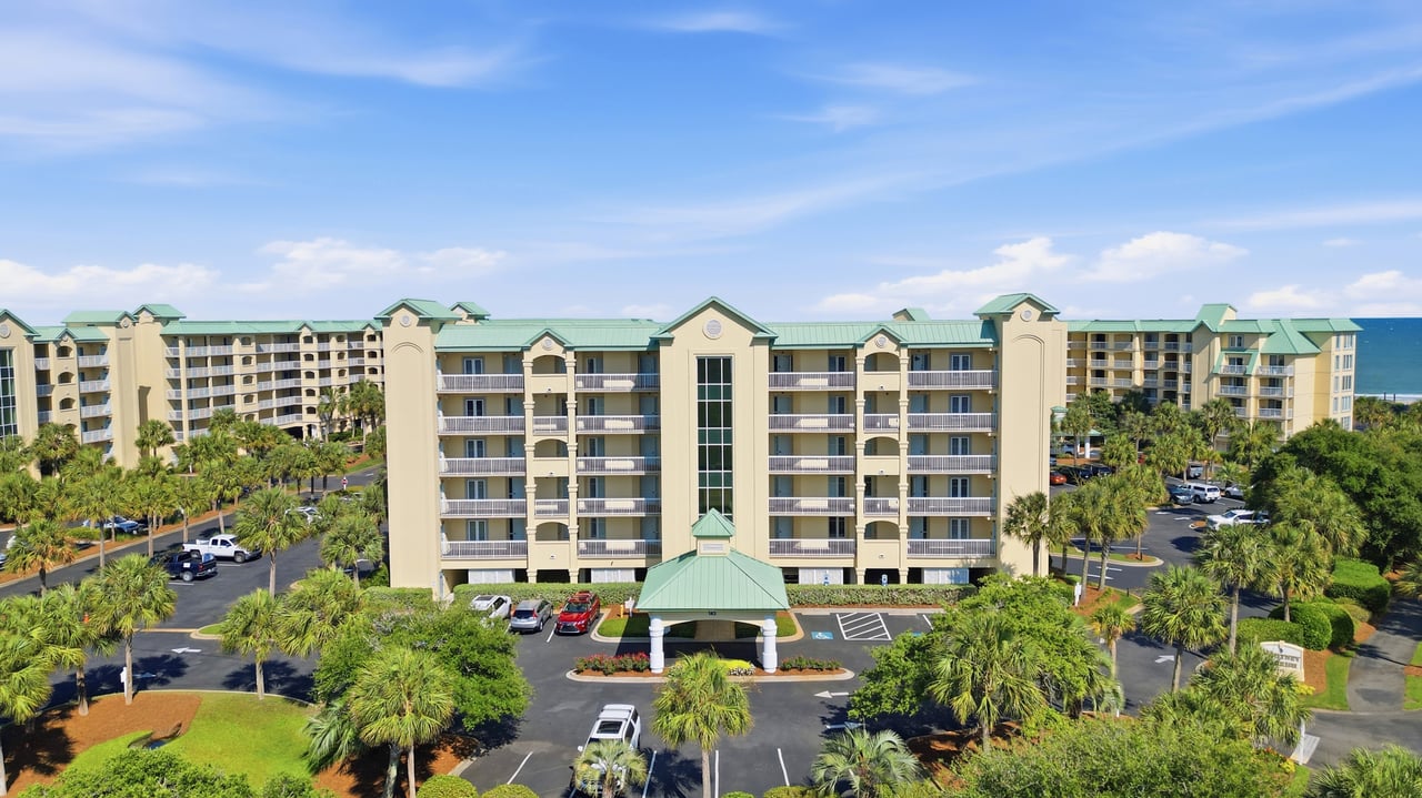 Coastal condominium complex with palm-lined grounds and ocean visible behind it.