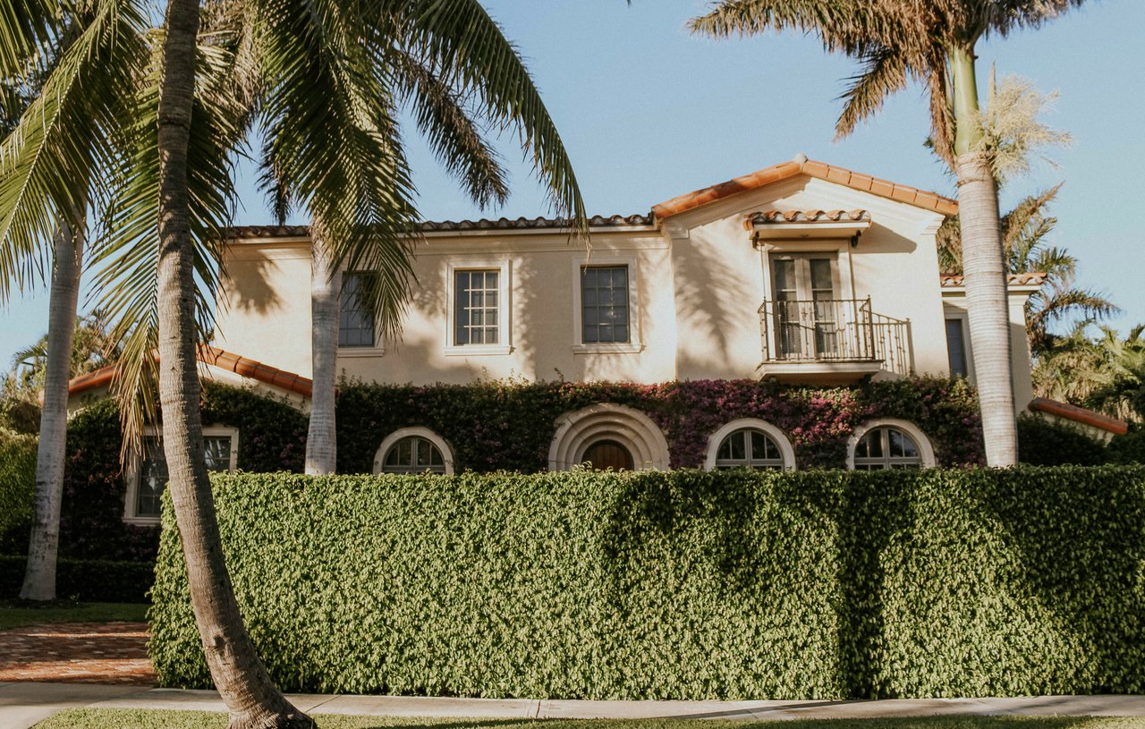 Two-story stucco home with a tiled roof, arched windows, balcony, and palm trees behind a tall hedge.