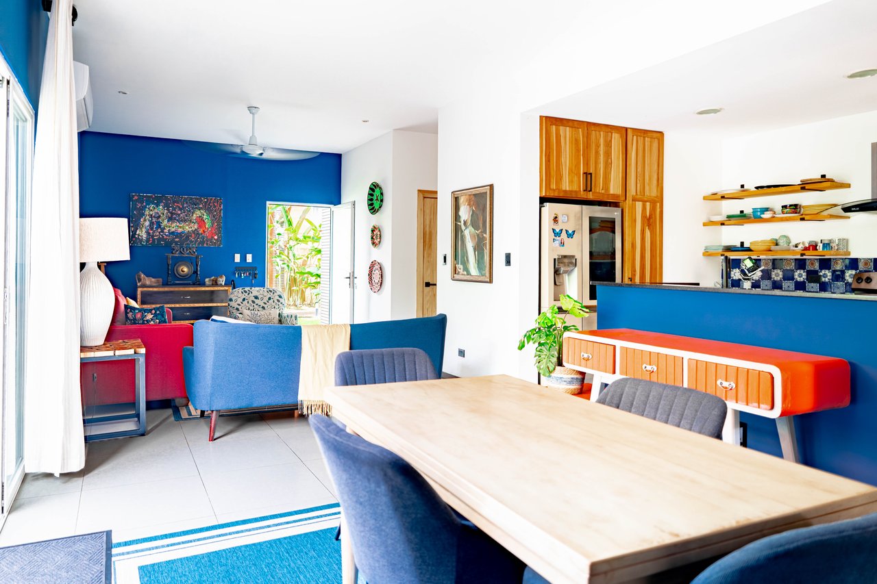 Open-plan living and dining area with blue accent walls, colorful furnishings, and a wood-cabinet kitchen.