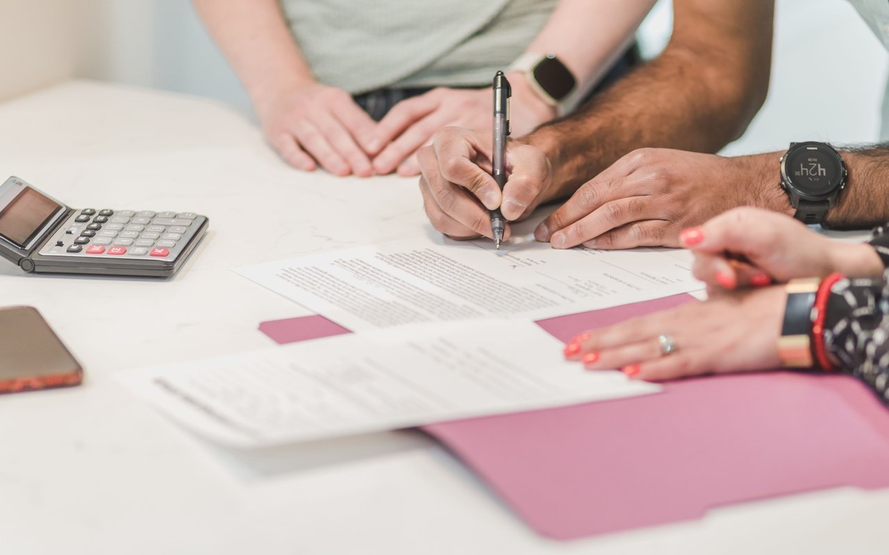 A person signs paperwork at a table beside a calculator and other documents.
