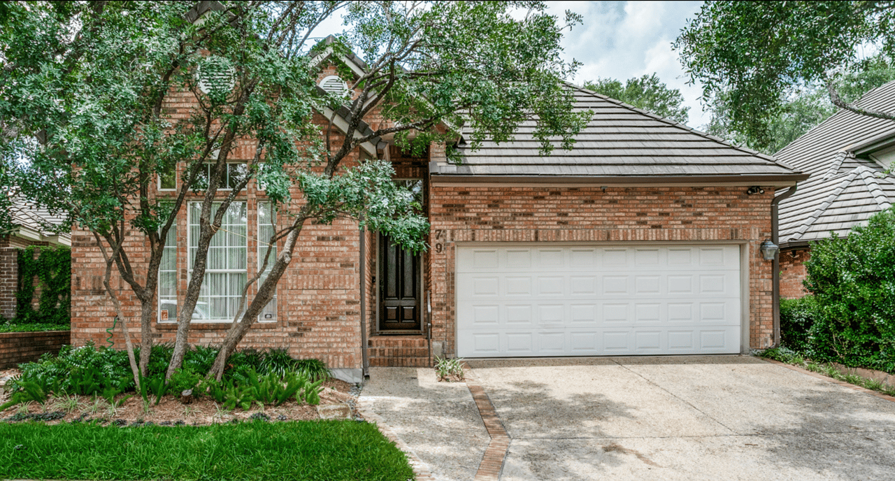 Brick one-story home exterior with white two-car garage, dark wood front door, and mature oak trees shading the front yard at 79 Longsford in Alamo Heights, San Antonio