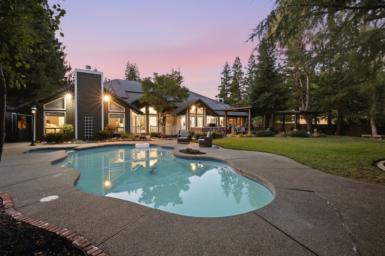 Modern home at dusk behind a curved swimming pool, with patio seating and a tree-lined yard.