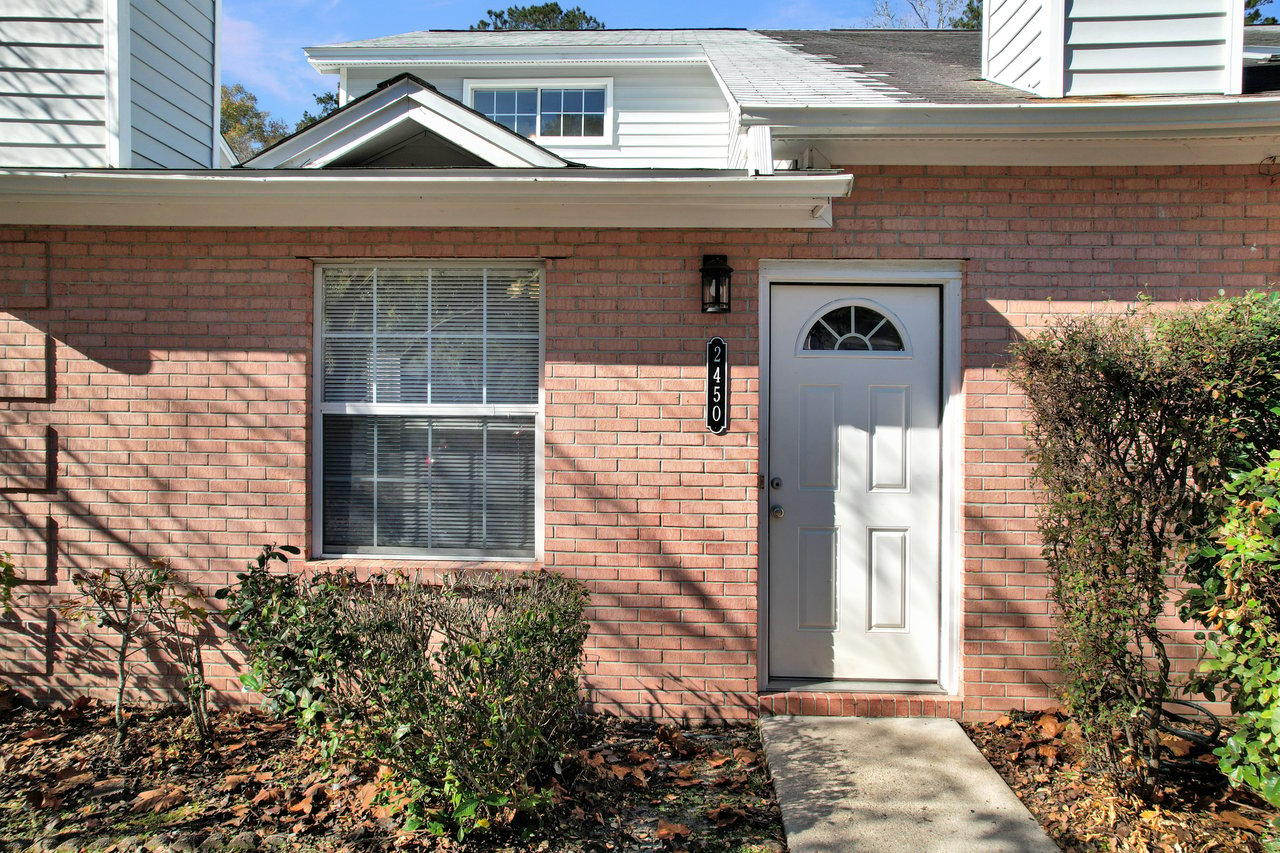 Brick house exterior with a white door and arched window. Sunlight casts shadows on the wall. Neat hedges line the path, conveying a calm, residential scene.