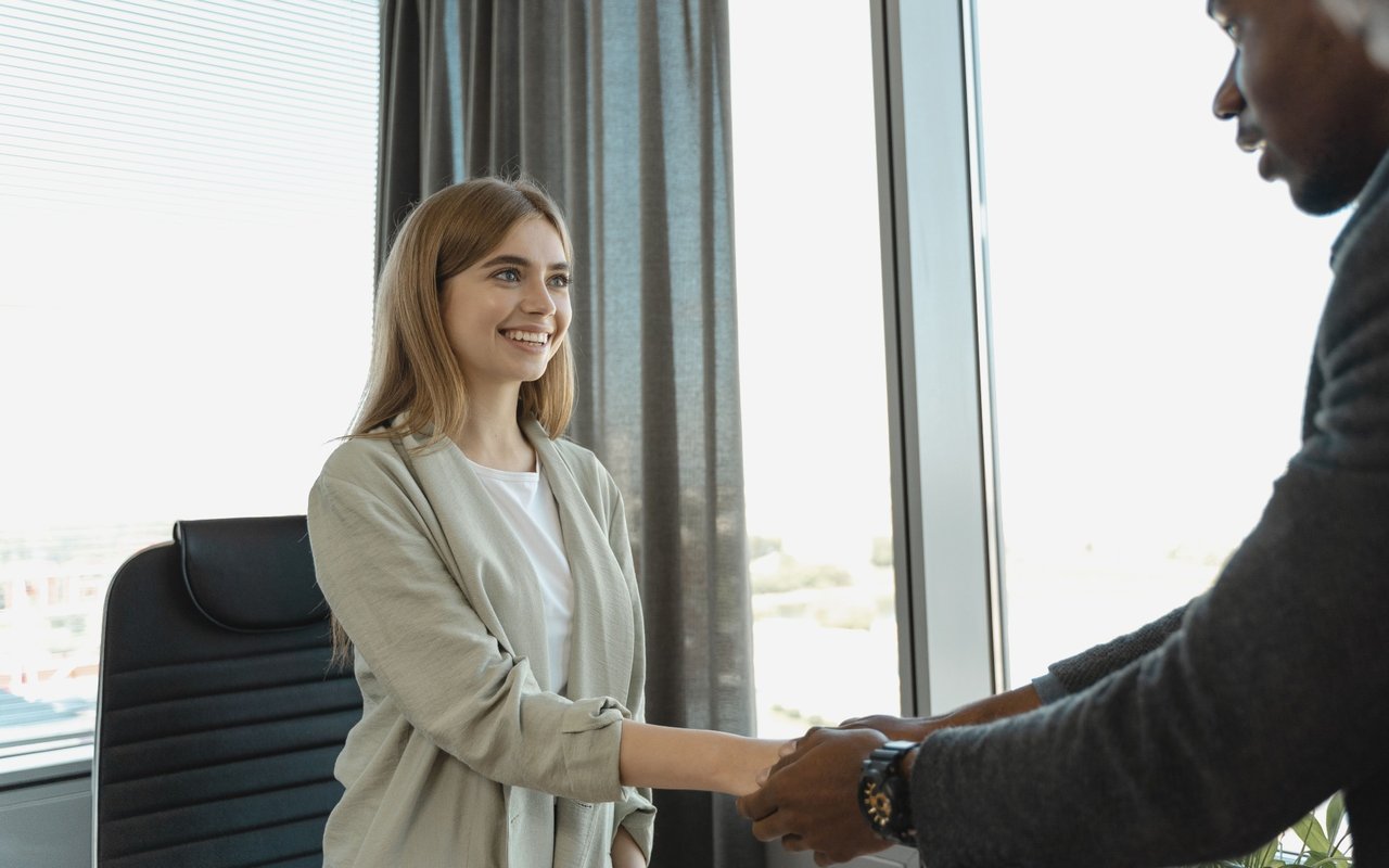 A smiling woman shakes hands with a man in a bright office.