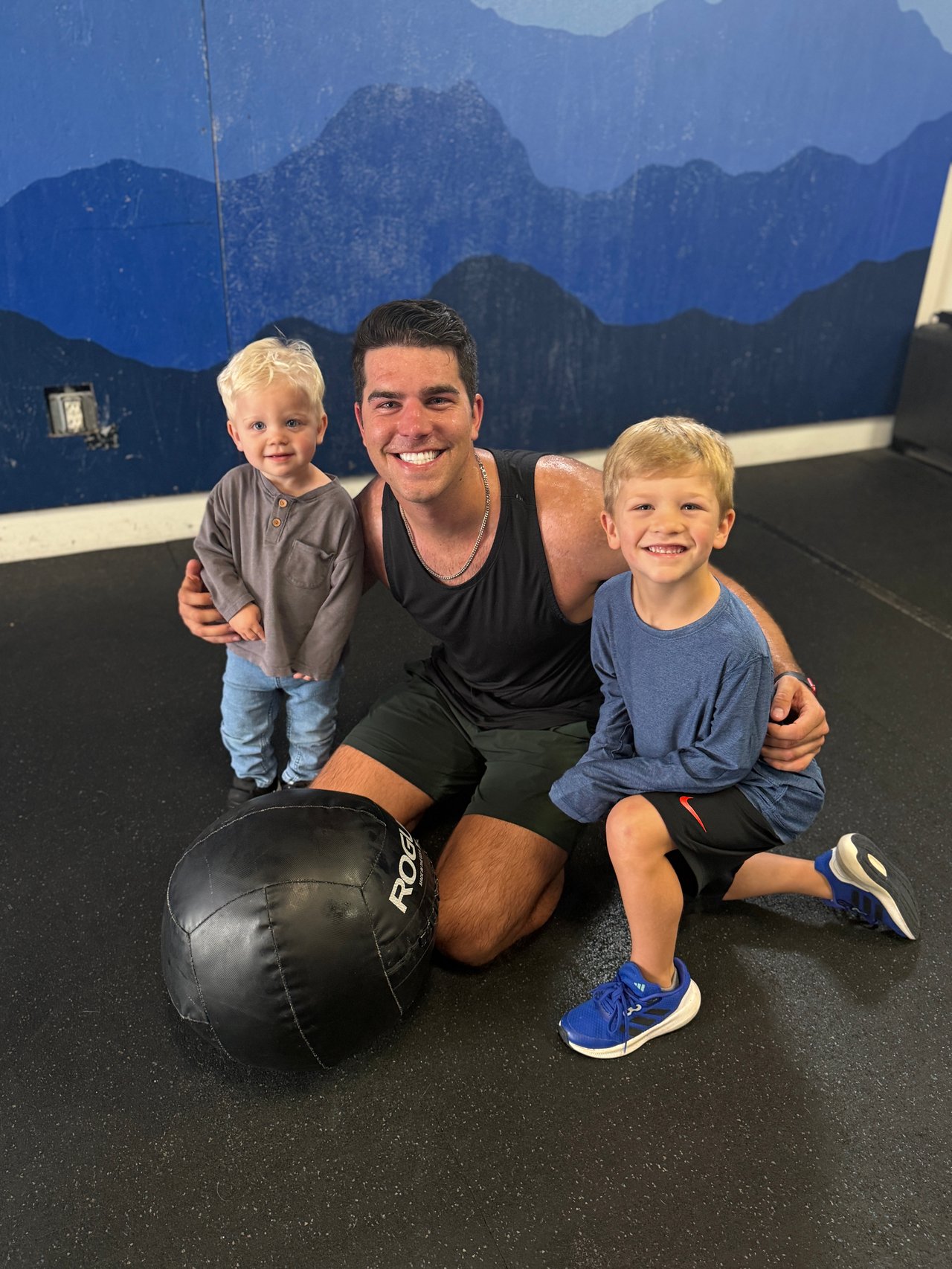A man kneels with two young boys beside a medicine ball in a gym.