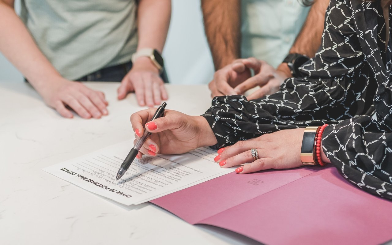 A woman points to a real estate purchase offer while signing it at a table.