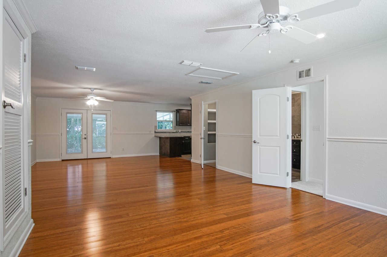 Spacious, empty living room with hardwood floors, ceiling fans, and French doors. Open kitchen area with dark cabinets visible, conveying an airy feel.