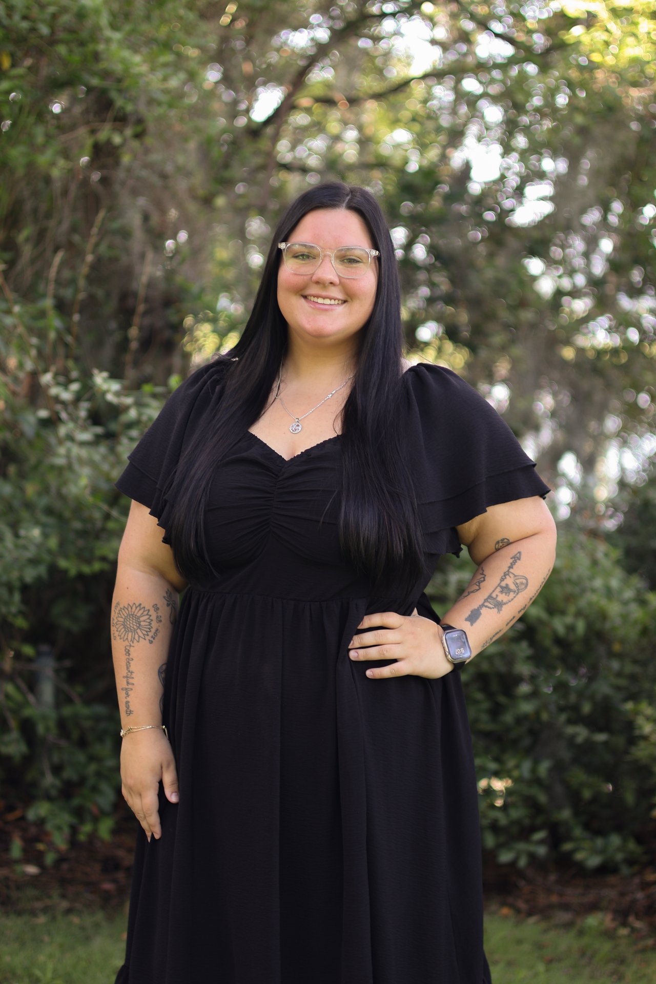 Emery Rook in a black dress, clear glasses, long dark hair, and tattooed arms.