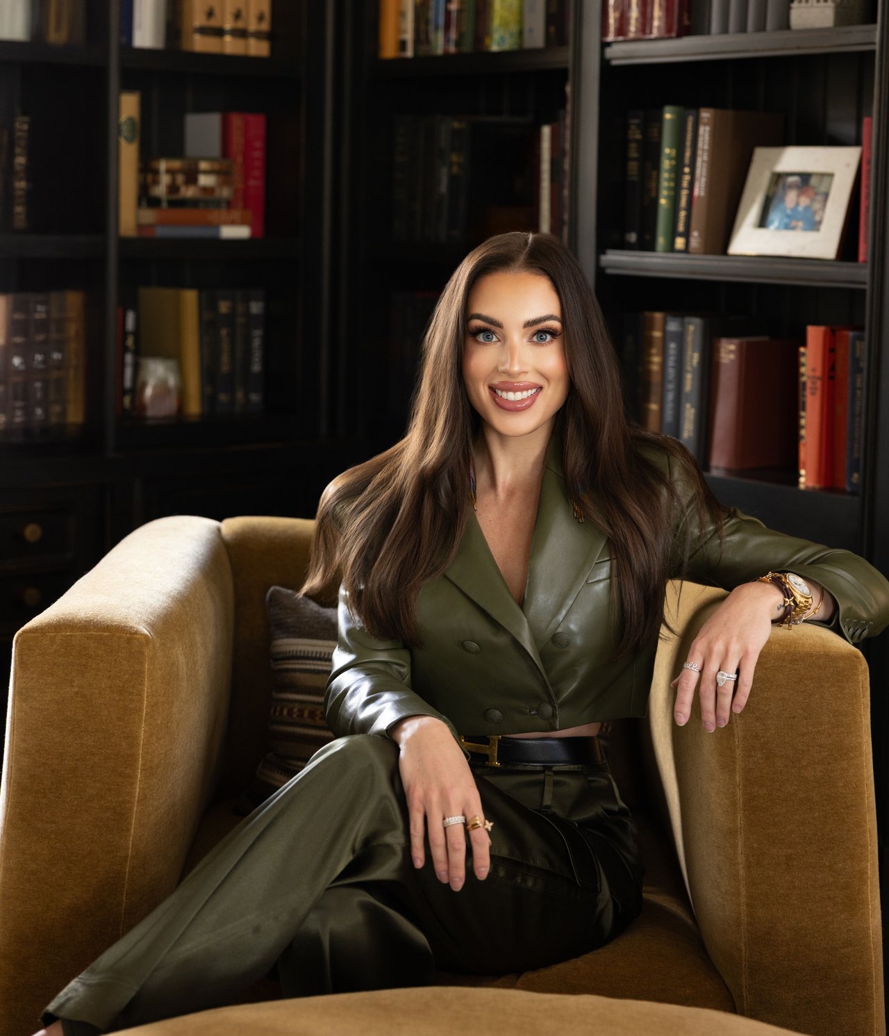 Woman in an olive-green suit seated in a tan armchair before bookshelves.