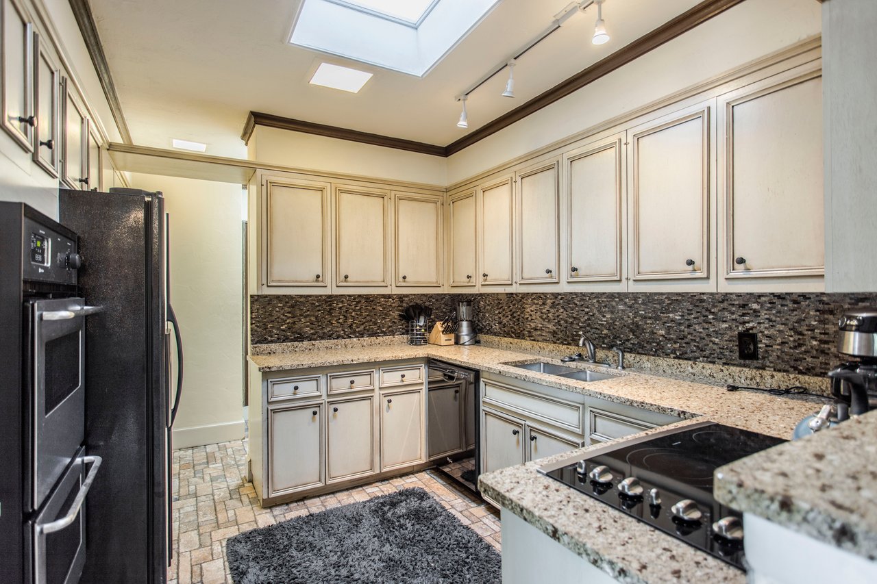 Kitchen with cream cabinets, granite counters, dark tile backsplash, black appliances, and a skylight.