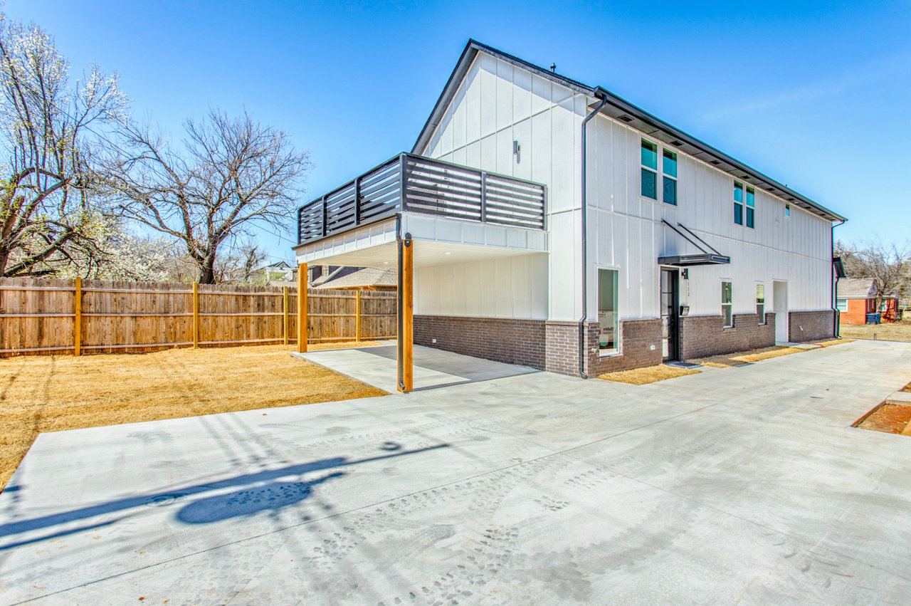 Modern white two-story house with a raised balcony, covered patio, and broad concrete driveway.