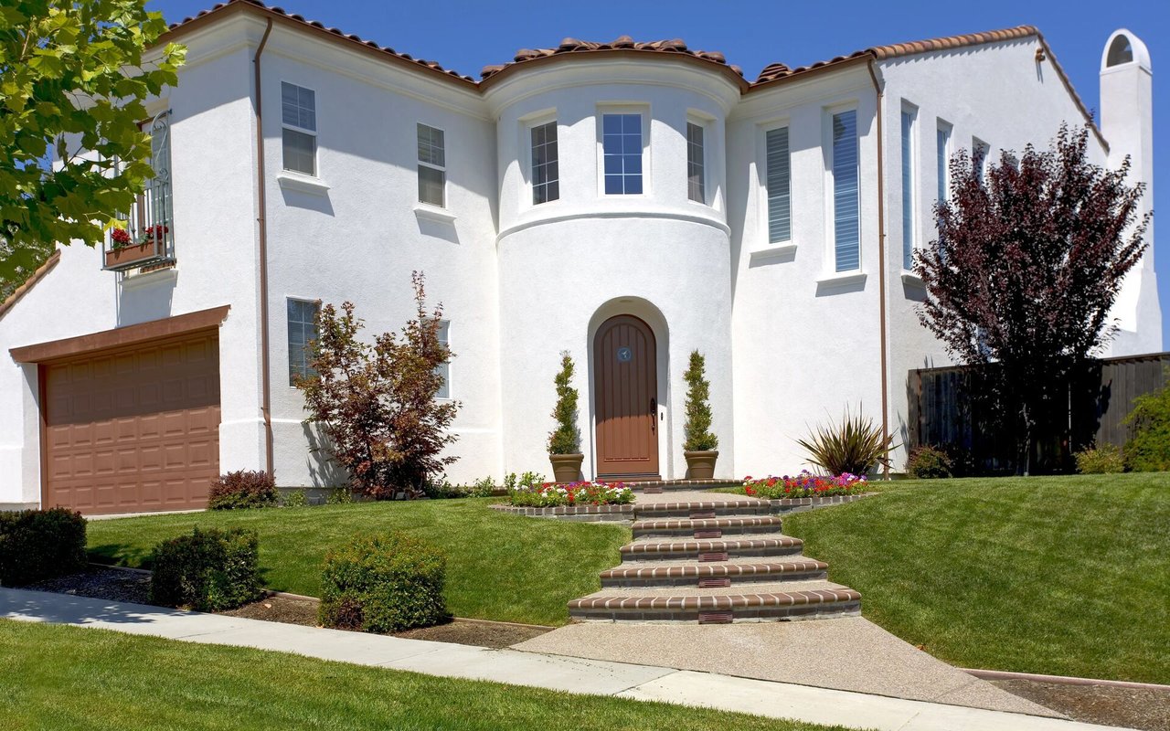 White two-story stucco house with an arched entry, curved brick steps, and a landscaped front lawn.