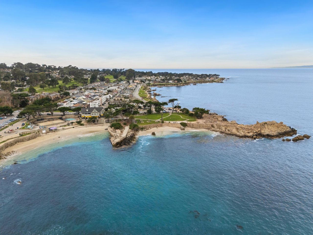 Aerial view of the Pacific Grove coastline with turquoise water, sandy cove beach, rocky points, and coastal homes near Lovers Point, Monterey County, California.