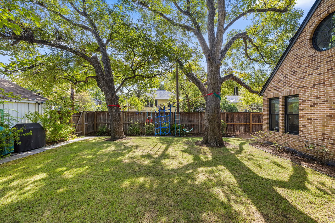 Fenced backyard with a grassy lawn, two large shade trees, and a blue climbing structure.