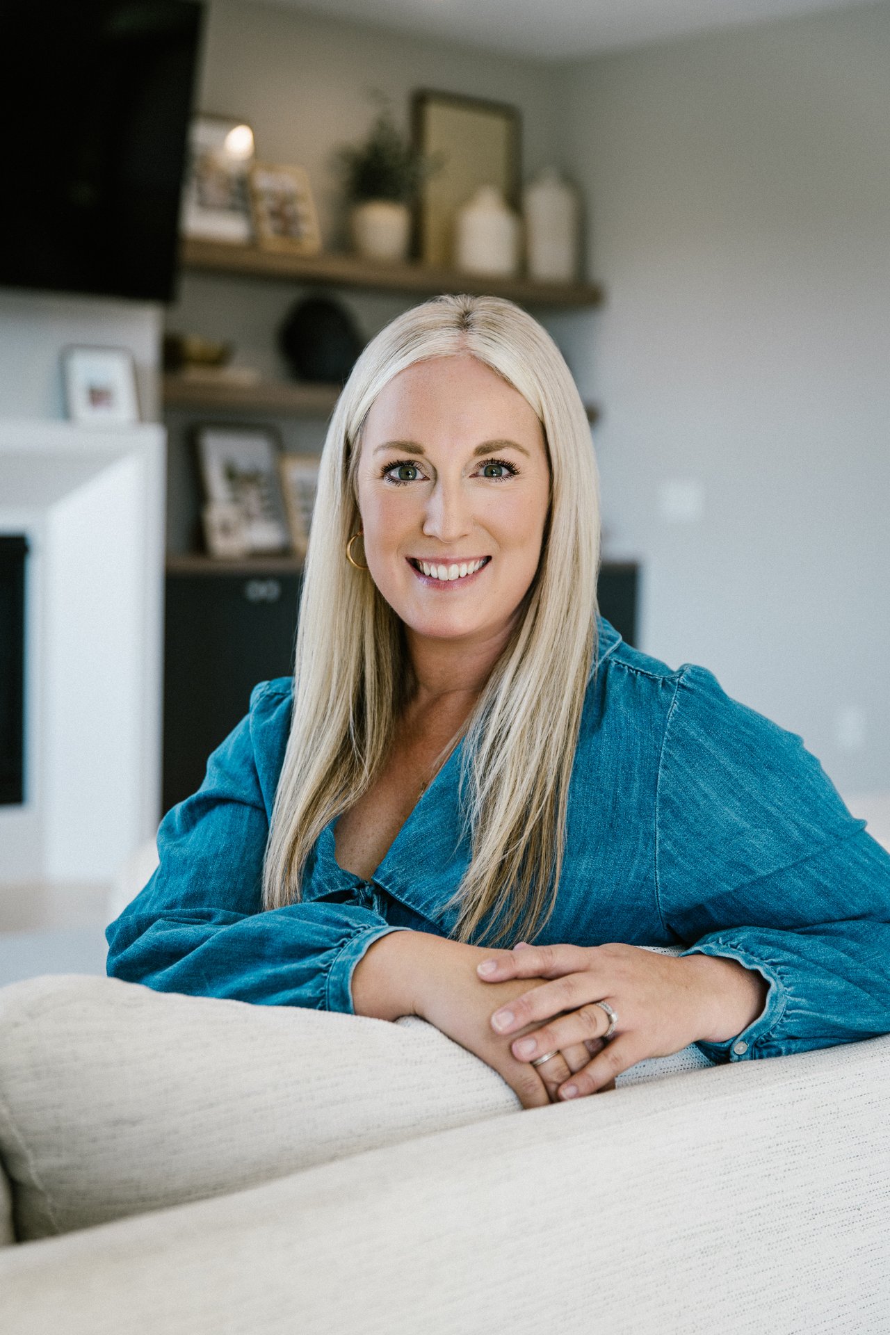 Hollie Engle smiling in a blue denim blouse while seated on a light-colored sofa.