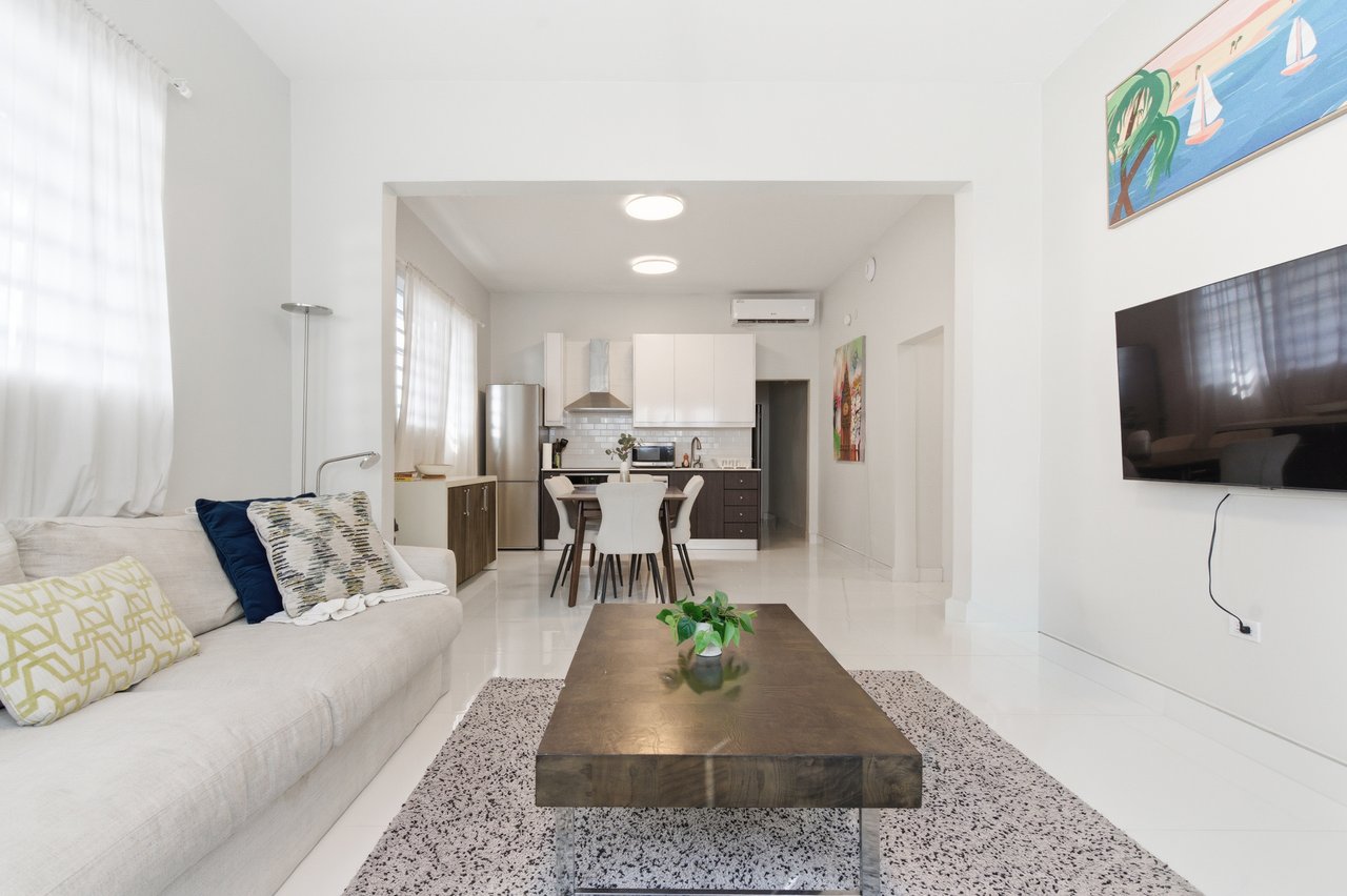Furnished living room with a cream sofa and dark wood coffee table, opening into a kitchen and dining area.