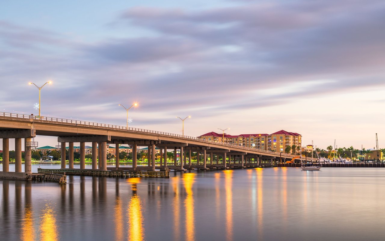 Bridge crossing a calm waterfront at dusk, with illuminated buildings and boats along the shore.
