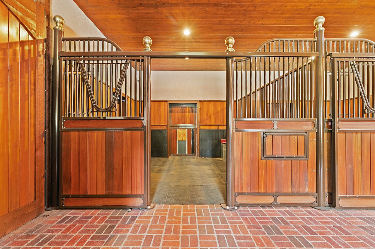 Equestrian barn interior with wooden stalls, metal gates, and a brick-paved entry.